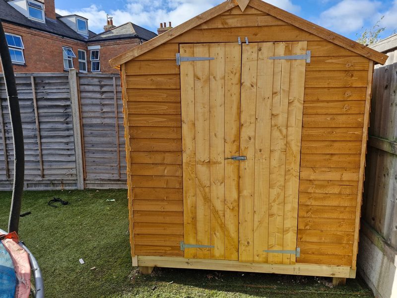 A timber apex-roofed garden shed with double doors, standing on a treated timber base on an artificial lawn.