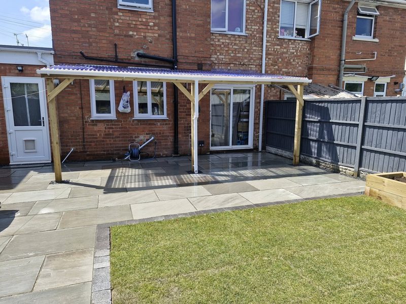 A timber veranda canopy with a clear corrugated roof, built over a sandstone patio at the back of a house.