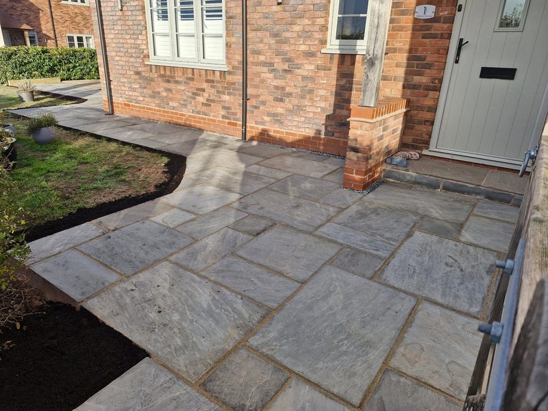 Wide grey sandstone paving laid around the front door and side of a new brick house, edged against a lawn.