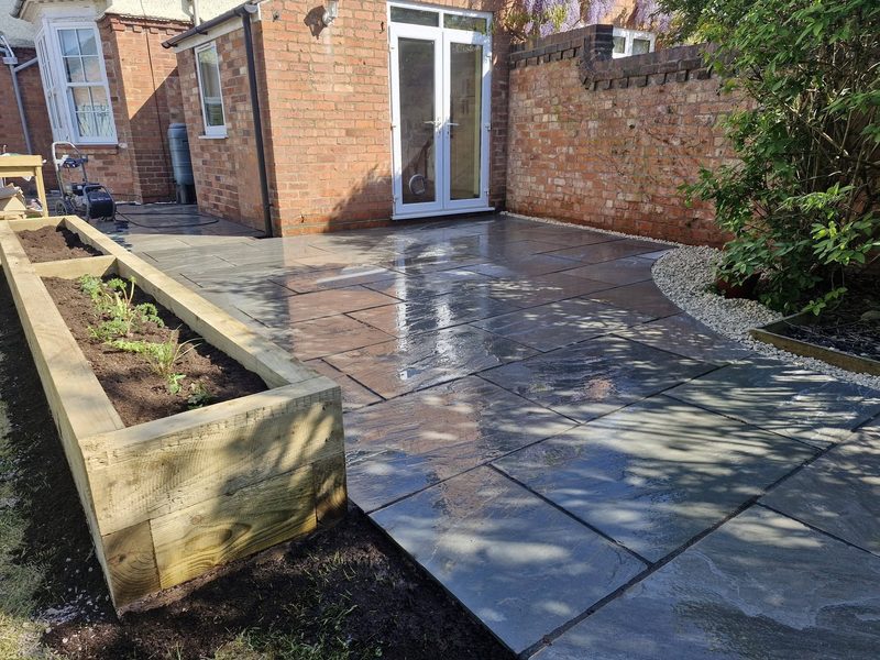A dark grey sandstone patio wrapping around a house, with timber raised beds planted up along one edge.
