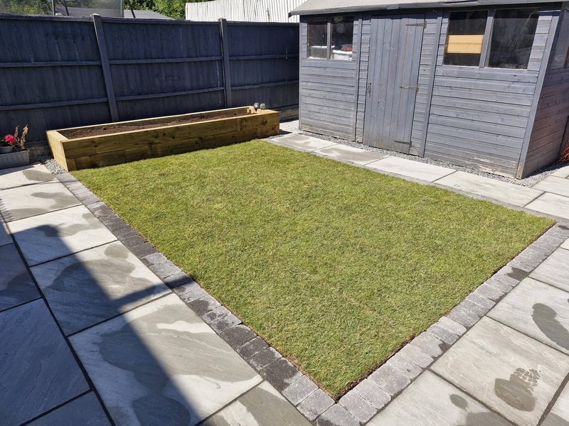 A new turf lawn edged with grey setts, bordered by sandstone paving, with a raised sleeper bed along the fence.