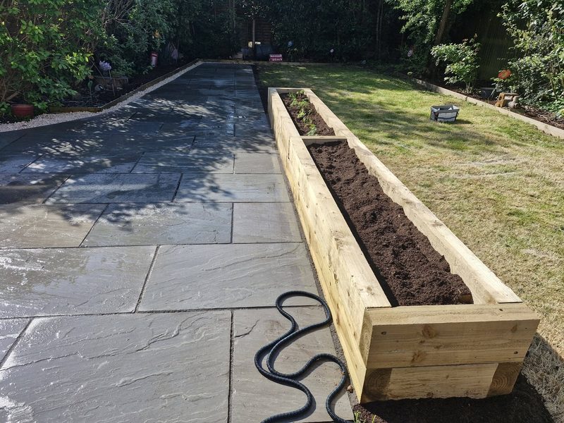 A wide sandstone patio and path with a long raised sleeper bed filled with fresh soil alongside a lawn.