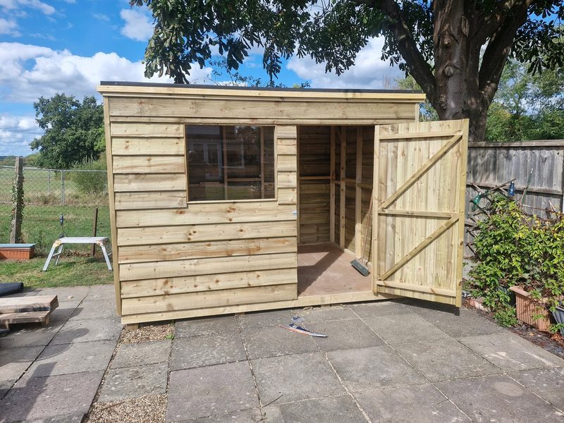 A newly built timber garden shed with a pent felt roof, window and open door, standing on a paved base.